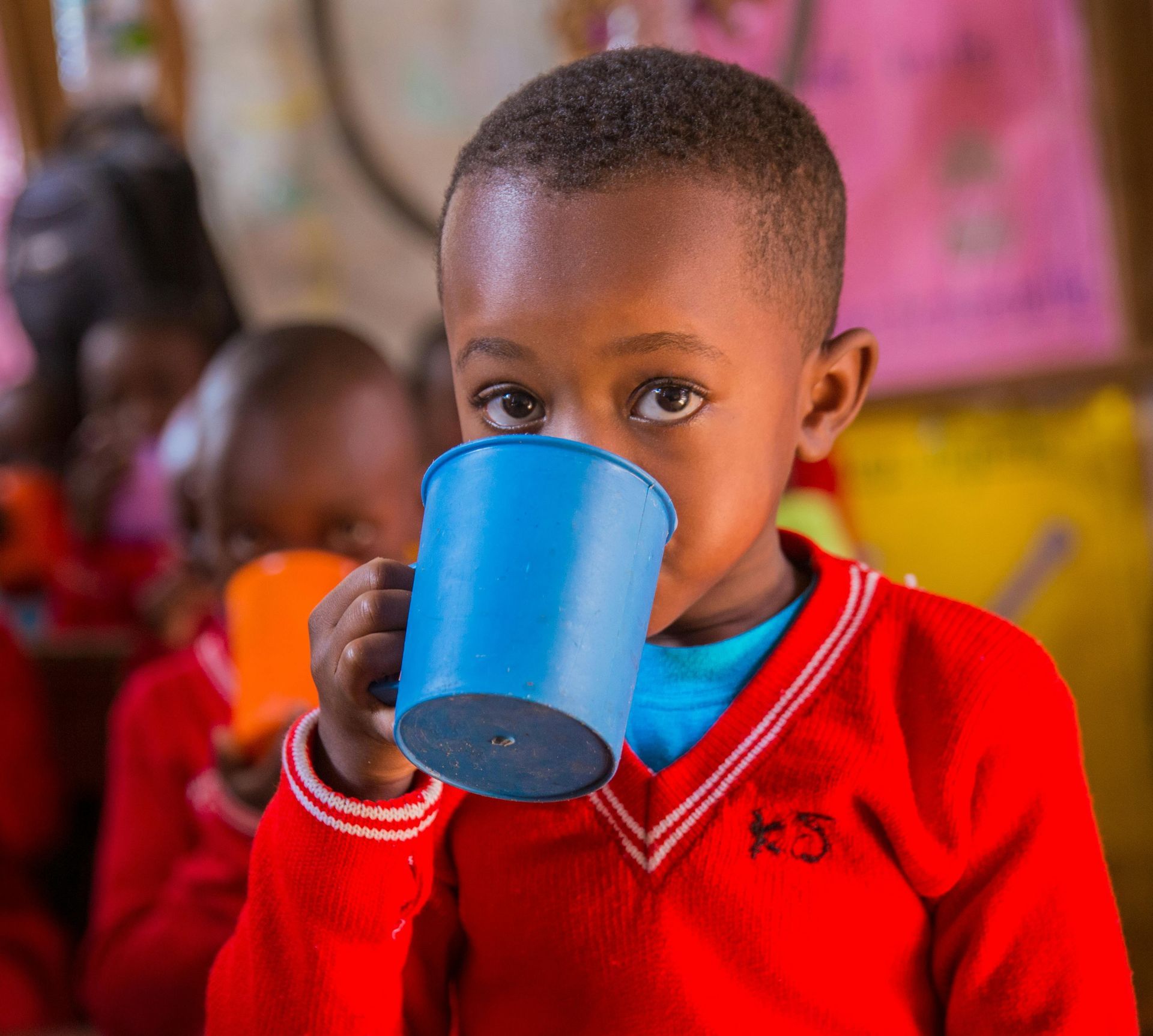 Boy in red sweater drinks from a blue cup; other children behind him.