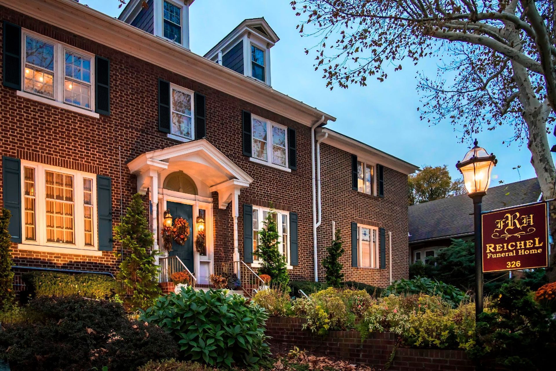 Brick inn at dusk with lit windows, white entrance, and a glowing sign among landscaped shrubs.