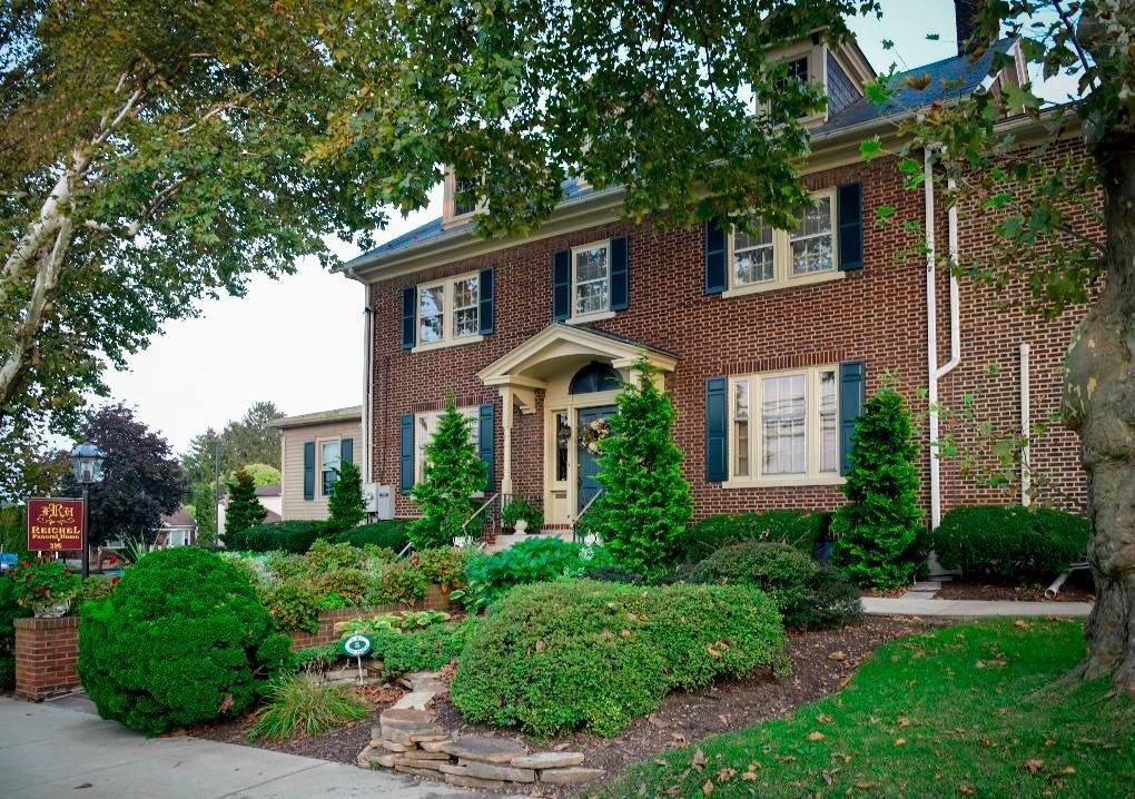 Brick two-story house with manicured front garden, white shutters, and a tree-shaded lawn