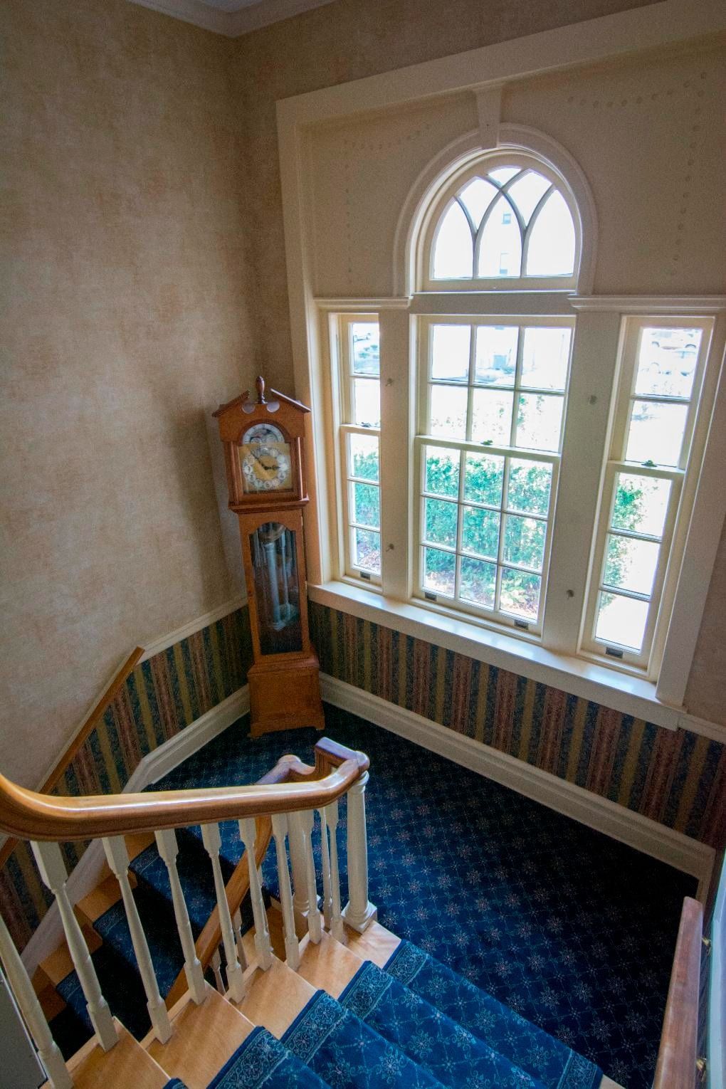 Staircase with blue carpet, tall grandfather clock, and large arched window in a bright hallway