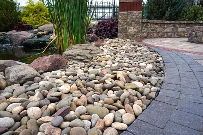 Curved garden path beside a bed of smooth river stones, with tall grasses and a stone wall