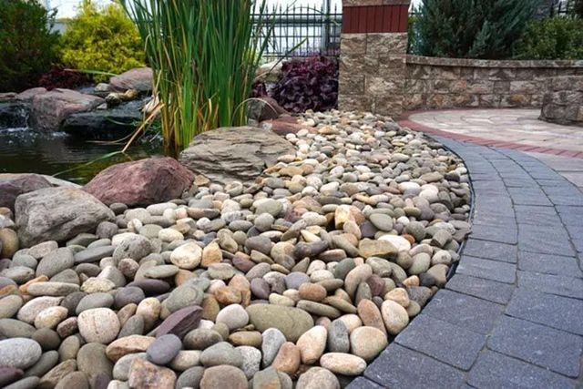 Curved garden path beside a bed of smooth river stones, with tall grasses and a stone wall