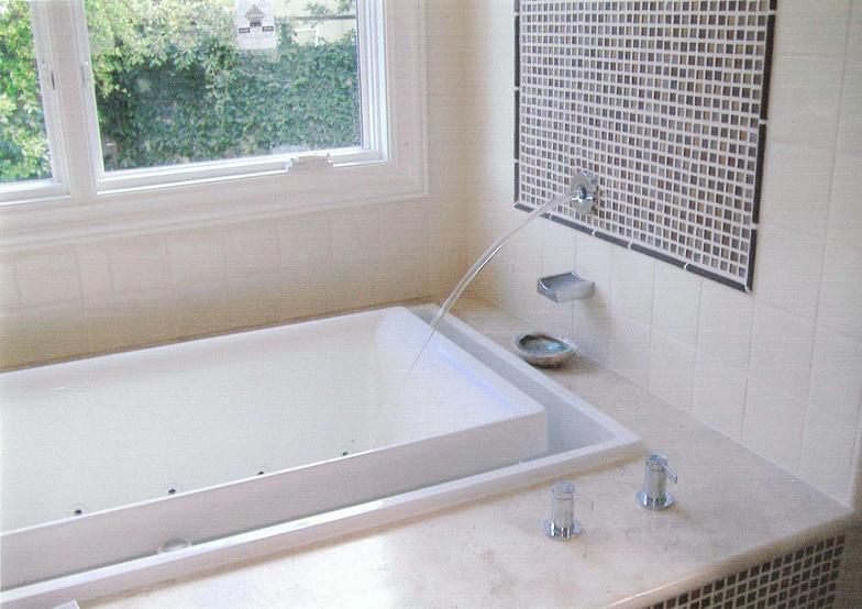 Modern bathroom with a rectangular white tub, window, and decorative tile wall feature. Water pours into the tub from a spout.