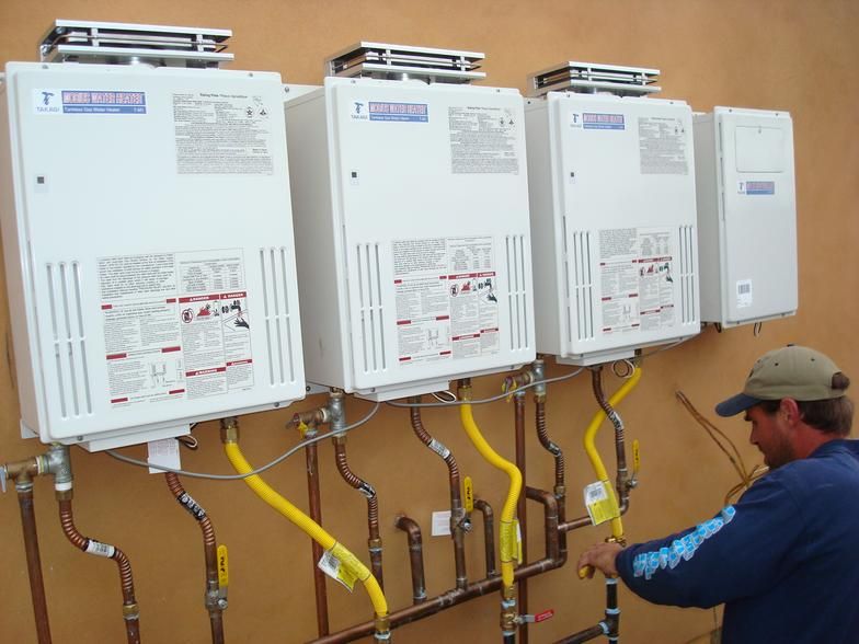 A person works on multiple wall-mounted water heaters with copper pipes and yellow gas lines outdoors against a brown wall.
