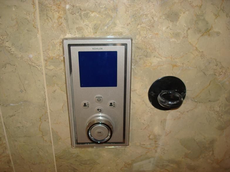 Silver shower control panel with a blue screen and a round knob, next to a black faucet on a marbled wall.