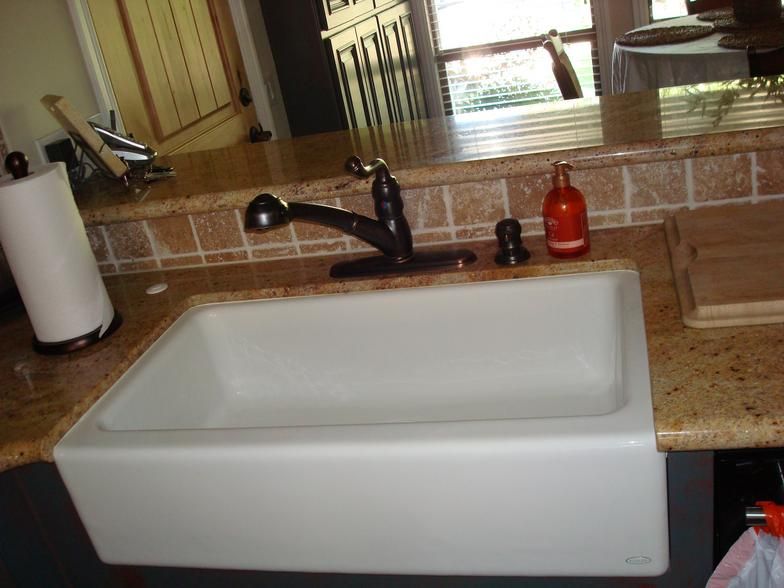 A white farmhouse sink with a black faucet in a kitchen. A paper towel holder and a bottle are visible.