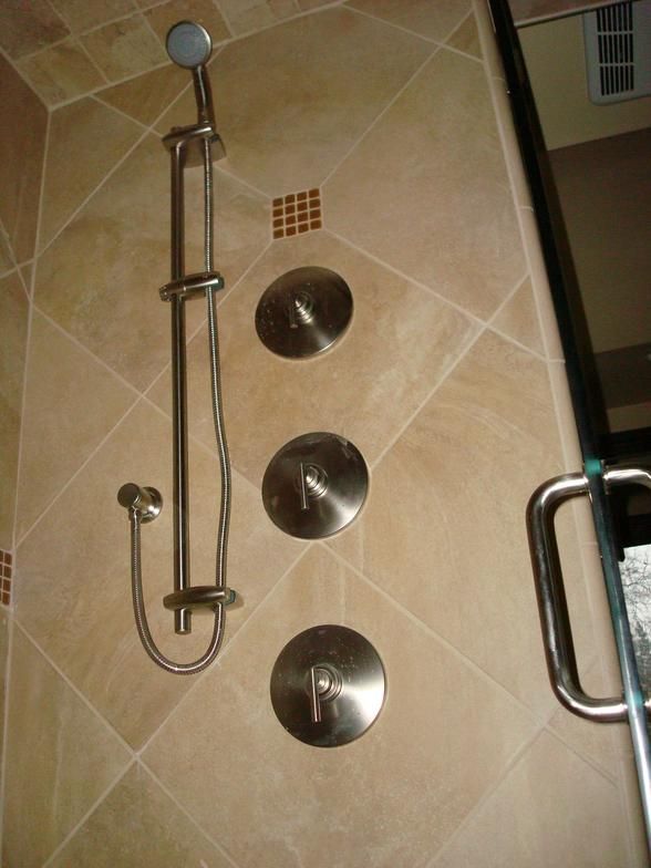 A shower interior with tan tile. Features a handheld showerhead, three water controls, and a glass door with a handle.