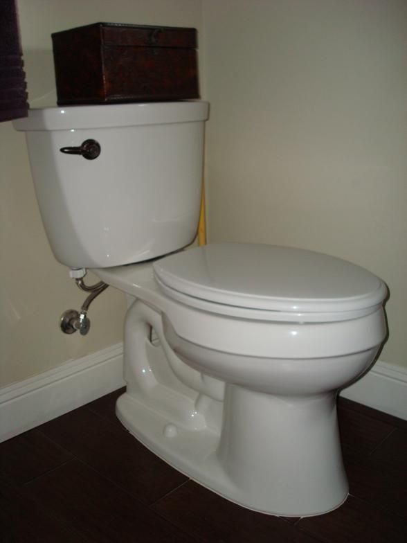 White toilet with a closed lid in a corner of a bathroom. A small brown box rests on the tank.