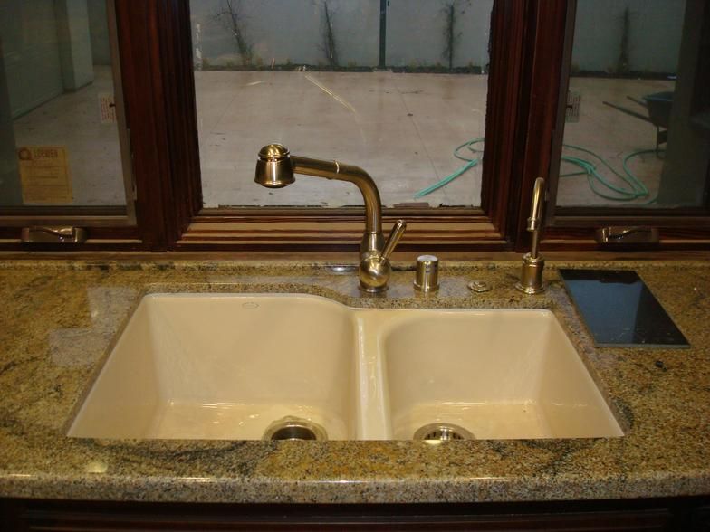 Kitchen sink with a gold faucet, two basins, and granite countertop in front of a window with brown trim.