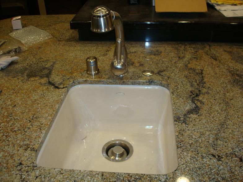 A kitchen sink made of white ceramic sits within a speckled granite countertop. A silver faucet and soap dispenser are present.