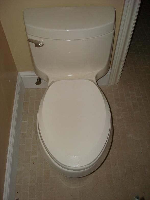 A white toilet with a closed lid, positioned in a bathroom setting. Beige walls and flooring are visible around the toilet.