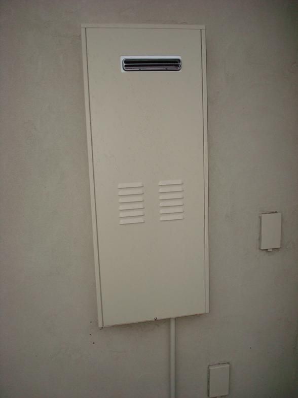 A rectangular, beige-colored utility box mounted on a light gray wall. It has a vent at the top and slits at the bottom.