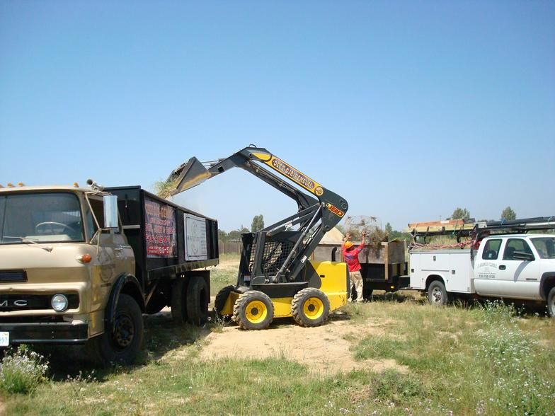 A yellow skid steer loader loads debris into a tan dump truck on a grassy field under a blue sky. Two other trucks are nearby.