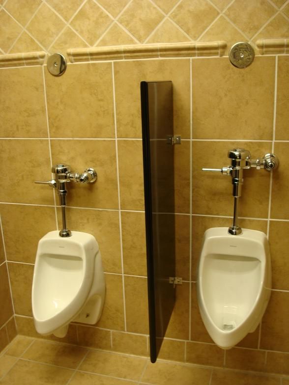 Two urinals in a public restroom with a dark partition between them. Beige tile walls and floors with silver flush handles.
