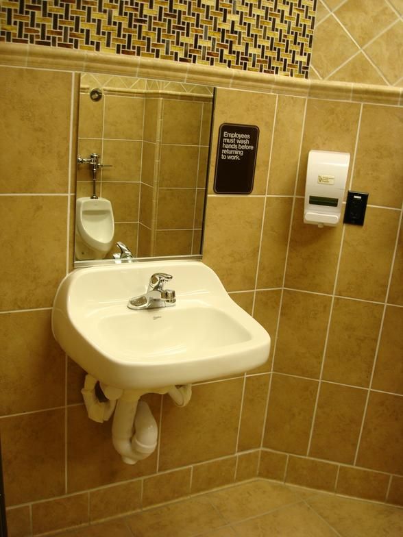 A public restroom with a wall-mounted sink and mirror; the floor and walls are tiled. A hand sanitizer dispenser is also on the wall.
