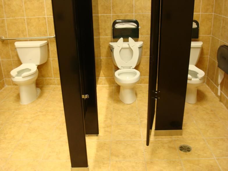 Public restroom with three toilets; tan tile walls and floor; black stall dividers.