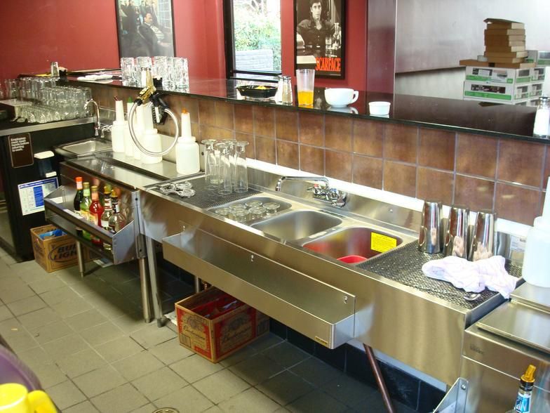 Stainless steel bar area with sinks, shelves of bottles, and glassware. Red walls and a bar top are visible.
