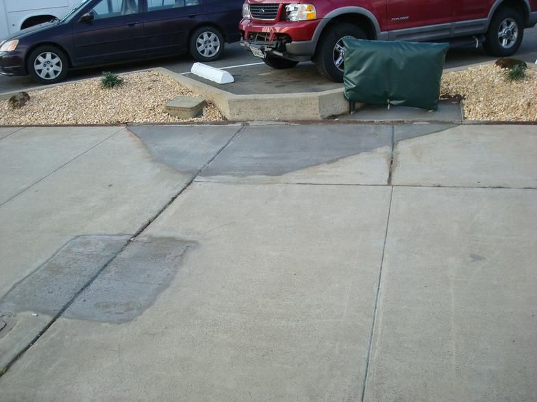 Cracked concrete sidewalk in front of parked cars and a bed of rocks and plants. Visible water stains.