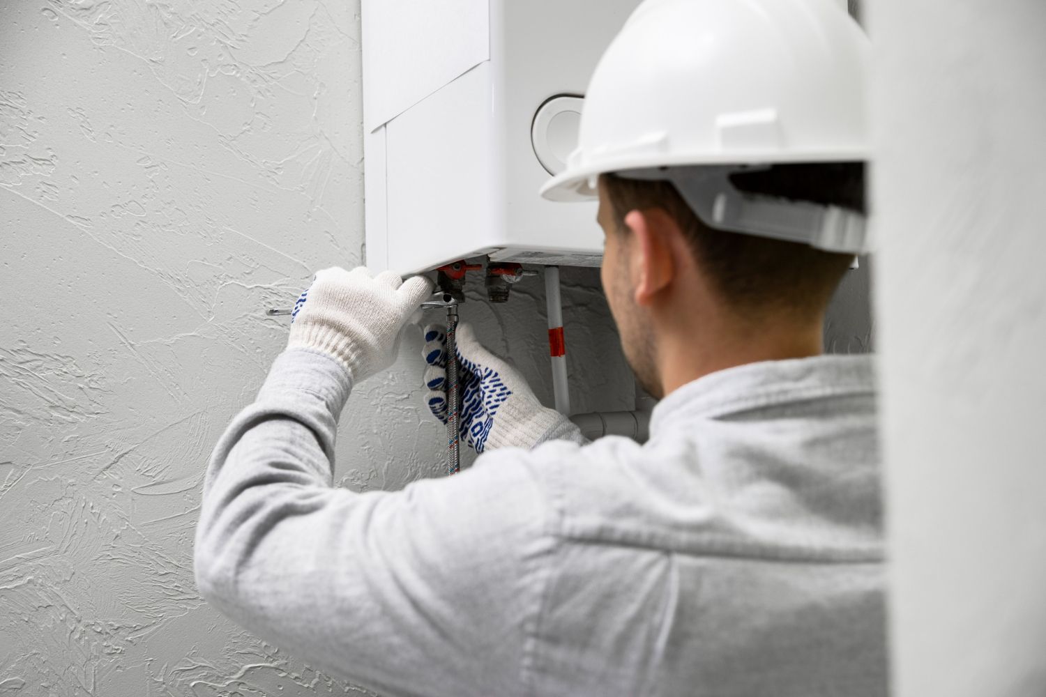 A person wearing a hard hat and gloves works on plumbing pipes connected to a white wall-mounted water heater.