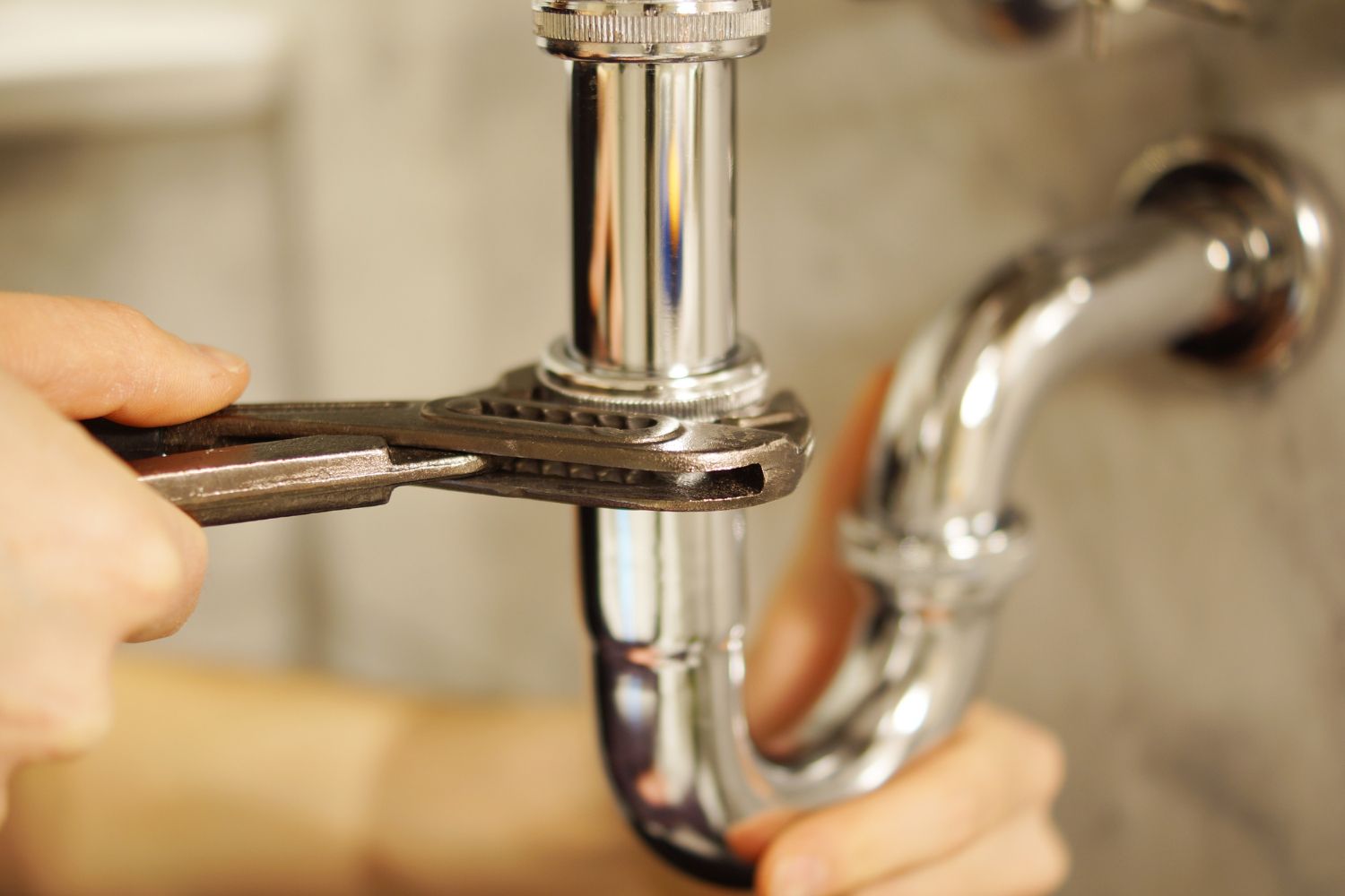 A person using a wrench to tighten a chrome pipe under a sink.