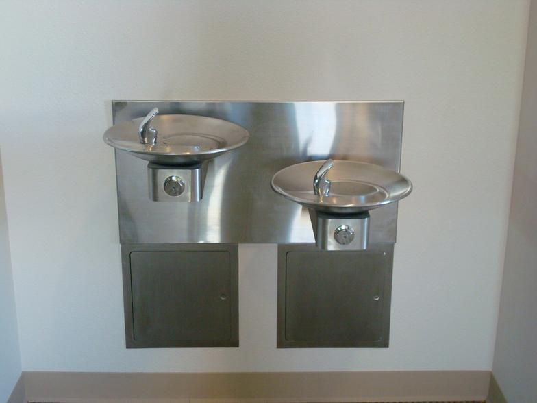 Two stainless steel water fountains mounted on a light-colored wall. Each has a circular basin and a button.