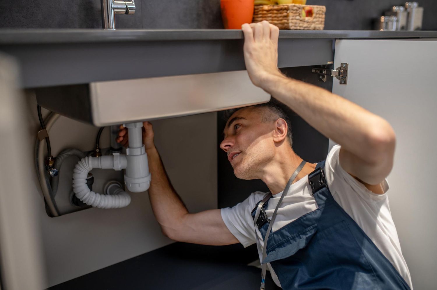 A plumber in overalls works under a sink, inspecting the pipes.