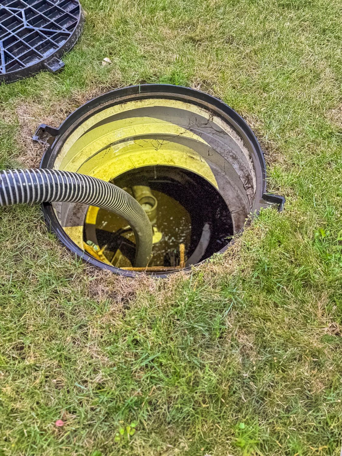 A septic tank being pumped on a grassy lawn; a large hose enters the open circular access point.