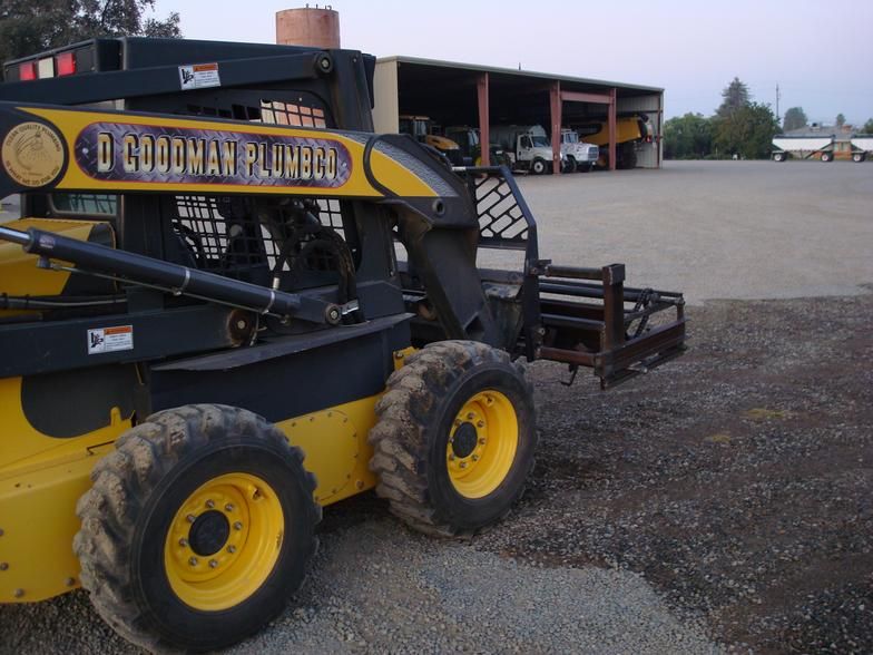 Yellow and black skid steer with fork attachment, parked on gravel, in front of a storage shed.