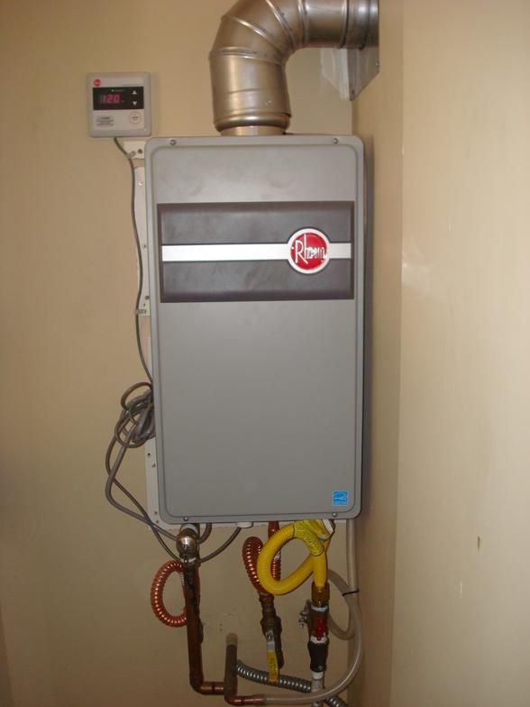 A Rheem tankless water heater mounted on a beige wall, with a silver exhaust pipe.  Plumbing and control panel are visible below.