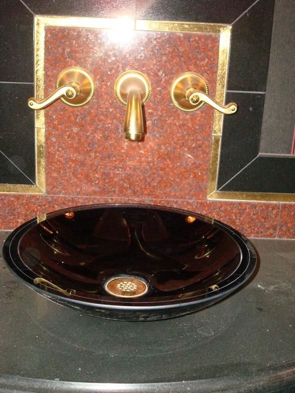 Oval glass bathroom sink with brass fixtures against a red and black tiled wall.