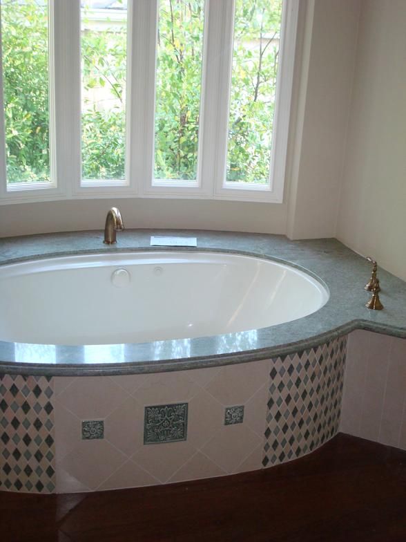 Oval bathtub with teal mosaic tile surround and granite countertop, near a window with a leafy green view.