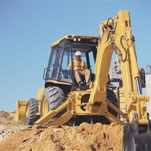 A yellow backhoe with a worker inside is on a dirt mound under a blue sky. The machine's bucket is scooping up soil.