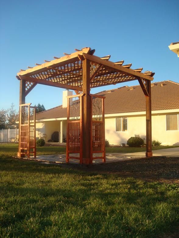 Wooden pergola in a yard, with latticework roof, surrounded by grass and a house in the background.