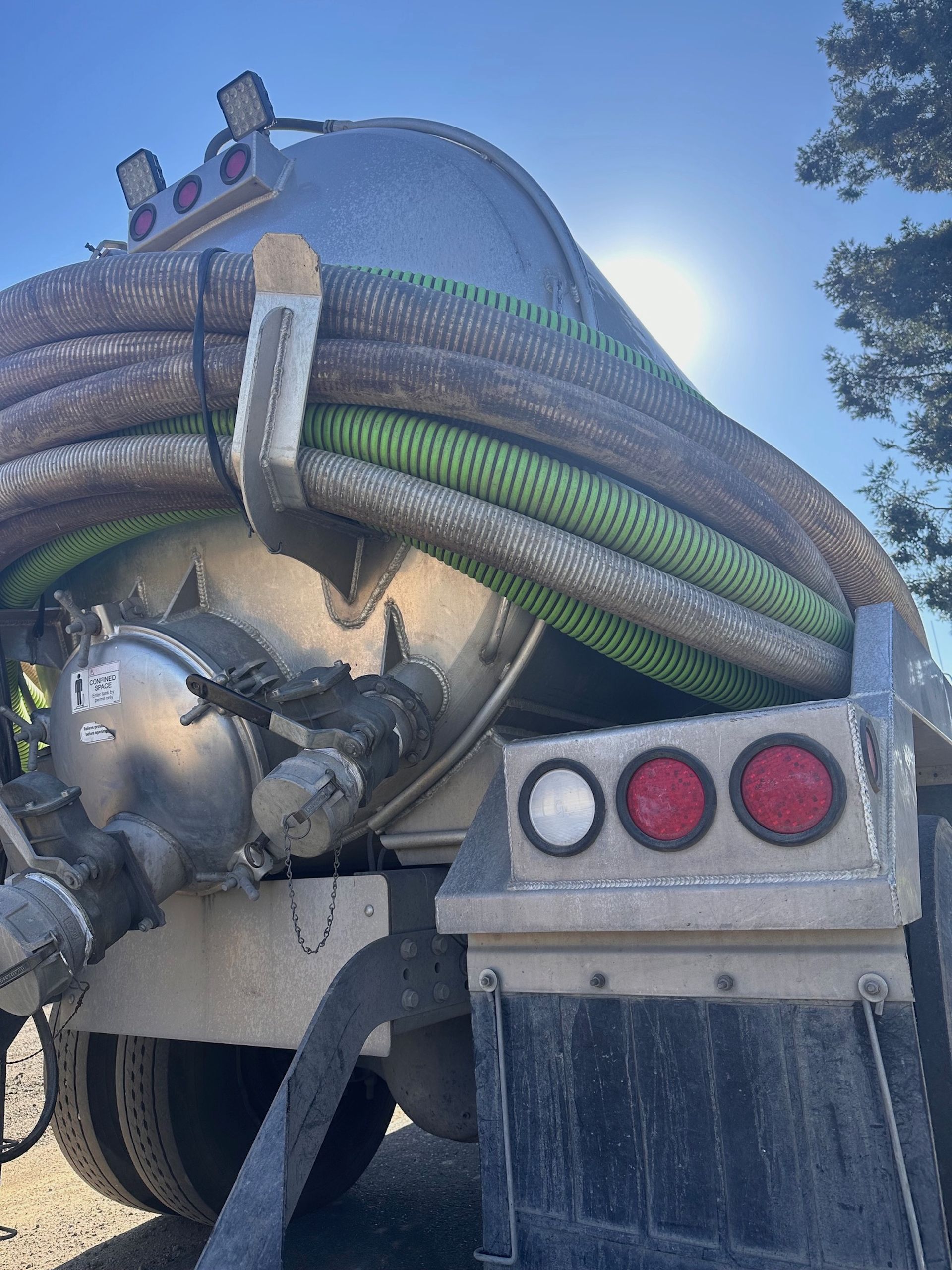 Rear view of a silver septic truck with hoses, bright sunlight, and black mud flaps.