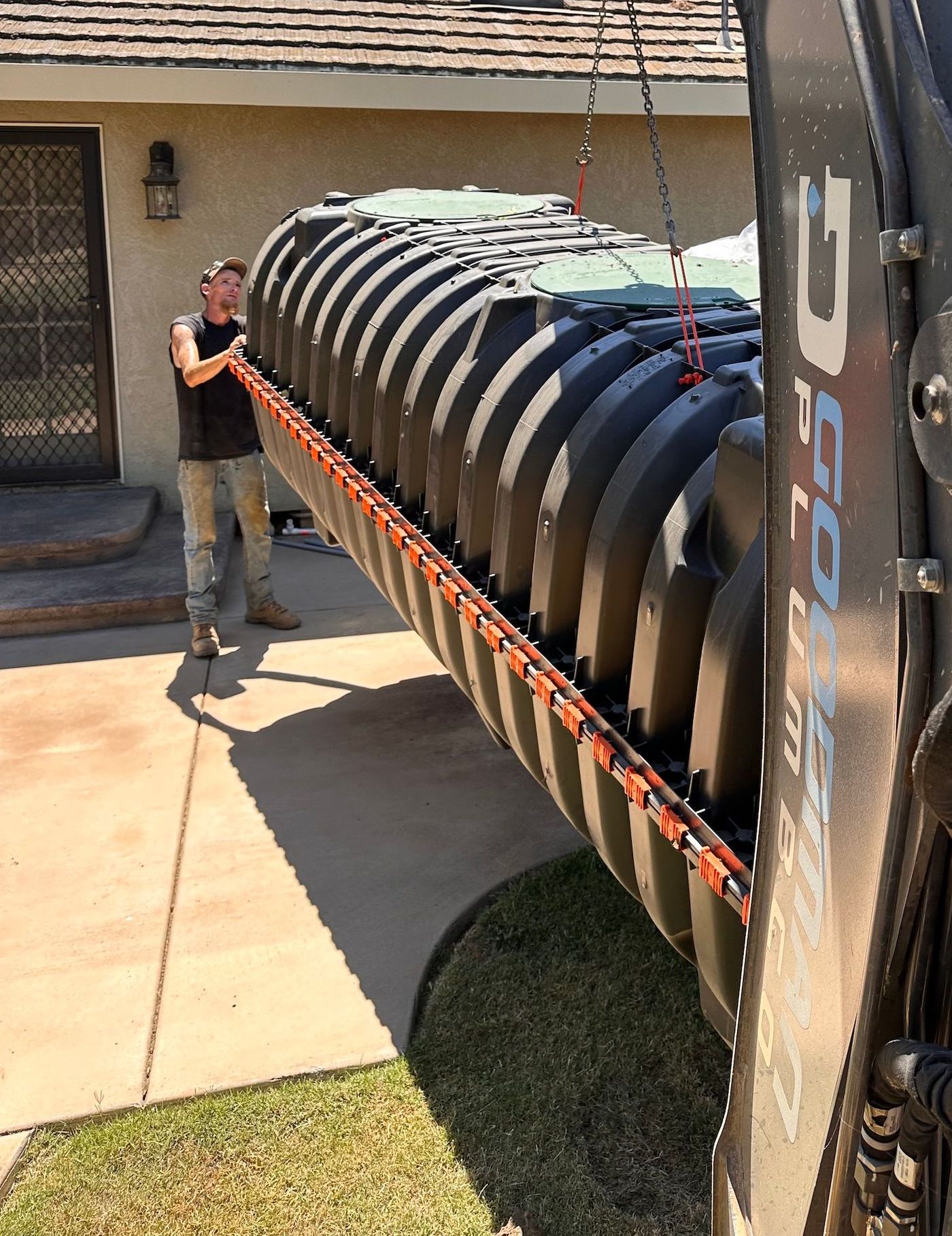 Man and machinery lift a large, dark sewage tank. They are outside a house with a door, on a paved area and lawn.