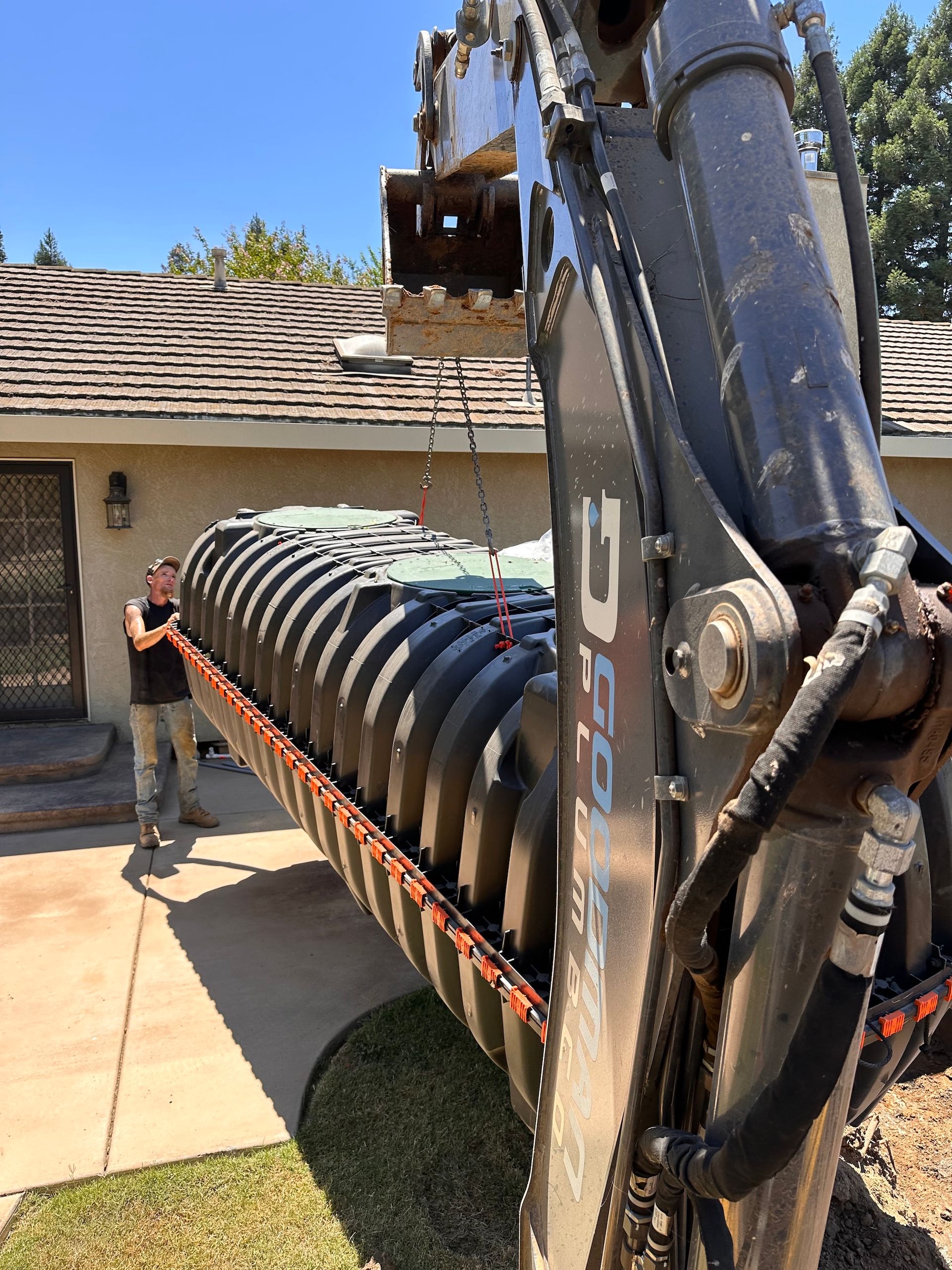 An excavator lifting a large, dark septic tank near a house.