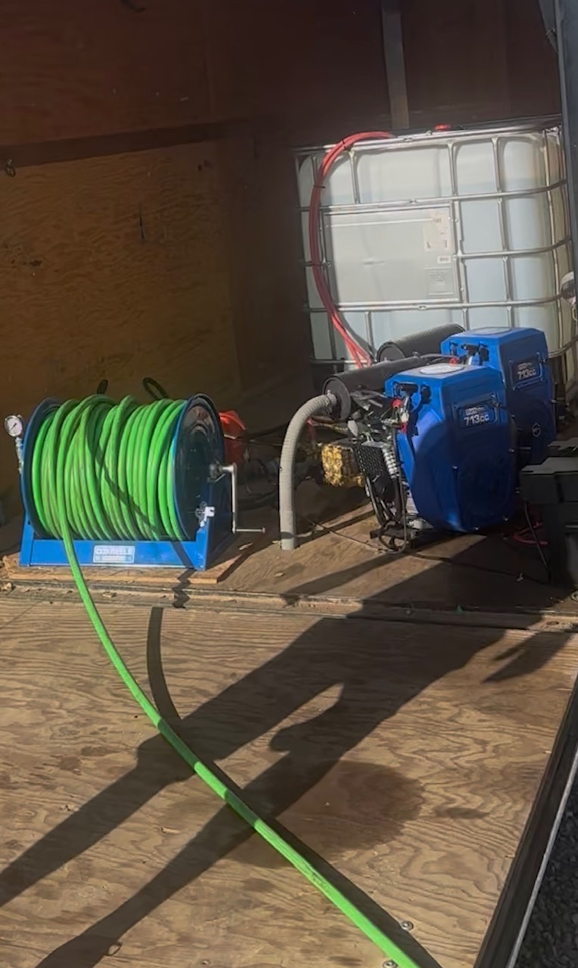 A pressure washing setup inside a trailer, featuring a green hose reel, a blue engine, and a white water tank.