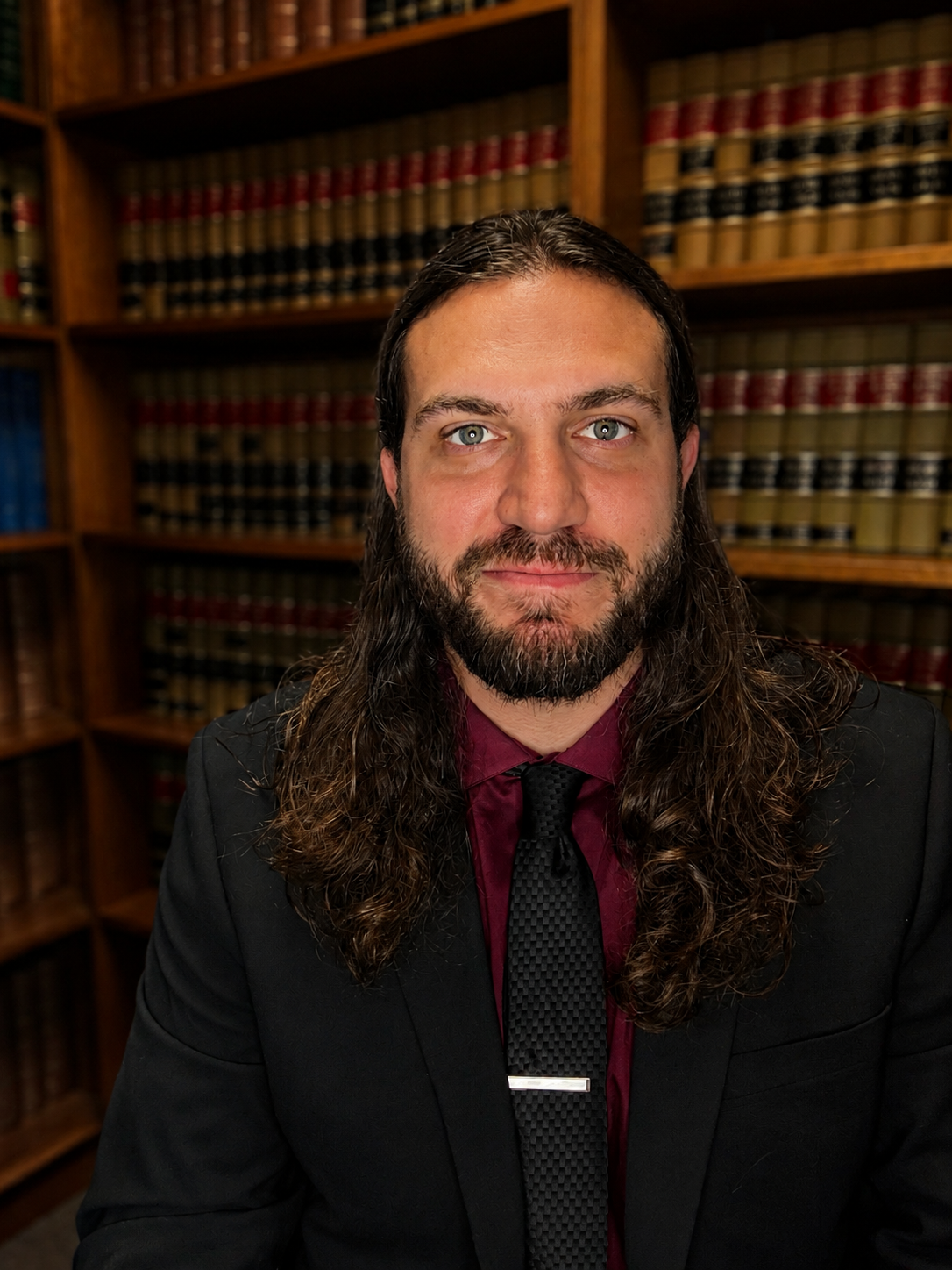 Brandon in a black suit and red shirt stands before bookshelves in a library, facing the camera.