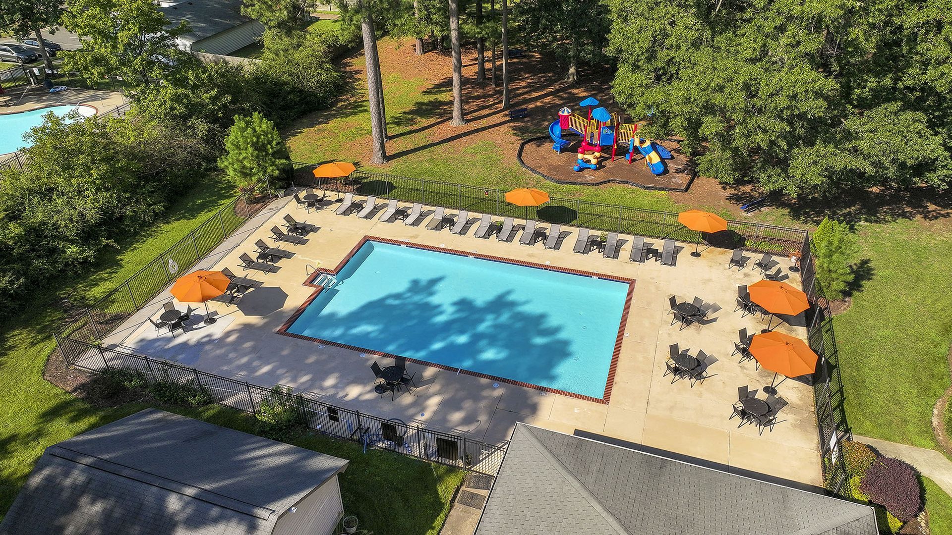 Aerial view of a fenced apartment pool area with lounge chairs and orange umbrellas; a playground is nearby at Chesapeake Pointe, offers  pet-friendly apartments in Chesapeake, VA