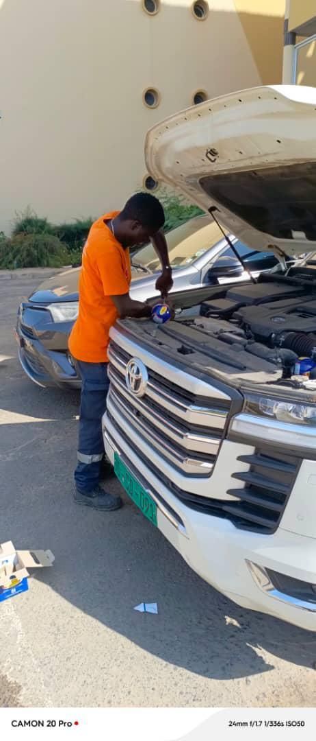 Un mécanicien en chemise orange travaille sur une voiture blanche avec son capot ouvert à l'extérieur.