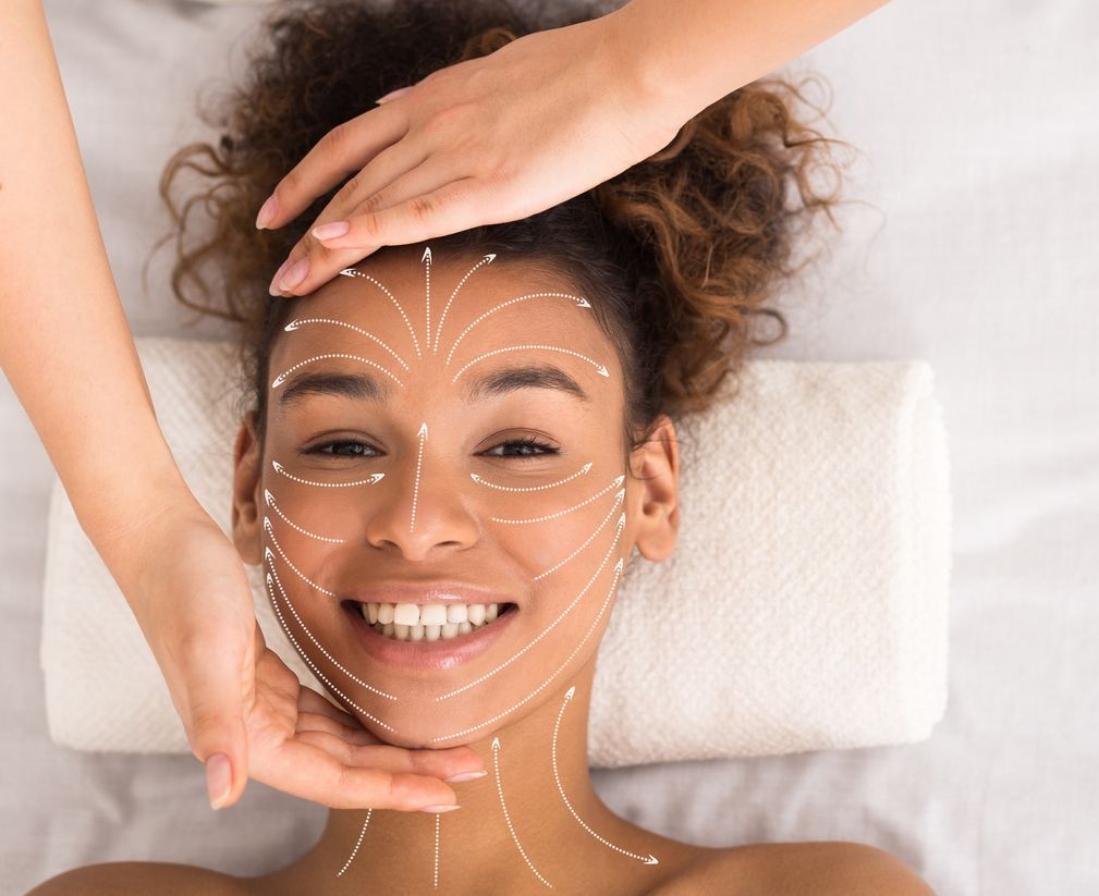 Smiling person receiving a facial massage, with white treatment lines on the face and a towel under the head.