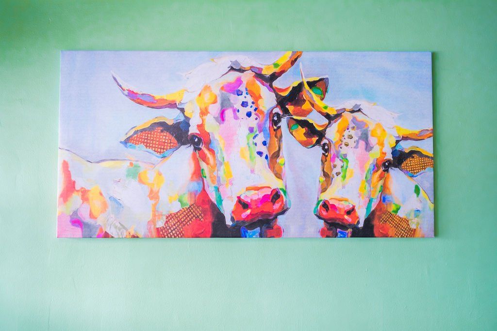Colorful abstract painting of two cows side-by-side, displayed on a solid light green wall.