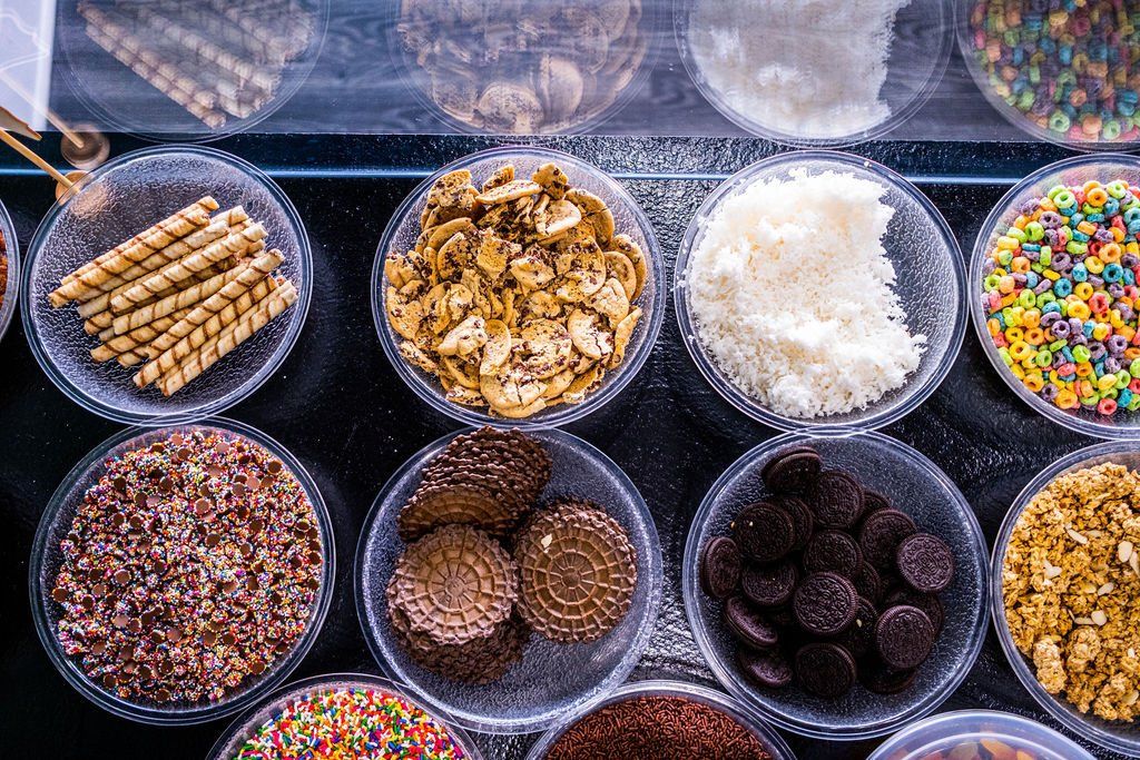 A variety of toppings for ice cream or yogurt, including cookies, cereal, and coconut, arranged in small clear bowls.