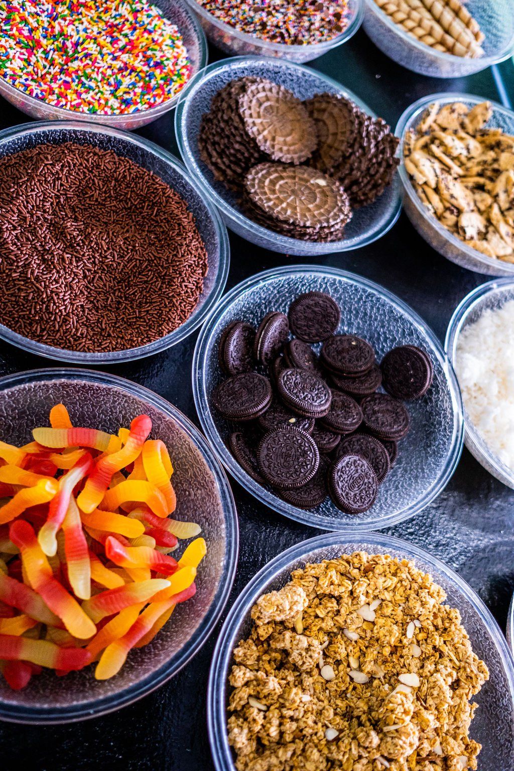 Various clear plastic bowls filled with toppings like gummy worms, chocolate sprinkles, cookies, and granola.