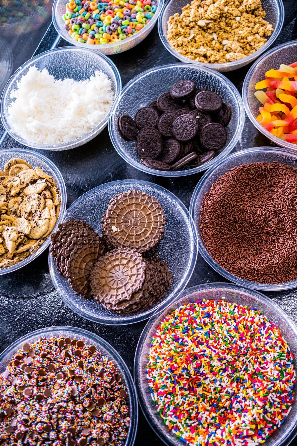 A high-angle view of multiple clear bowls filled with various ice cream toppings like sprinkles, cookies, and candy.