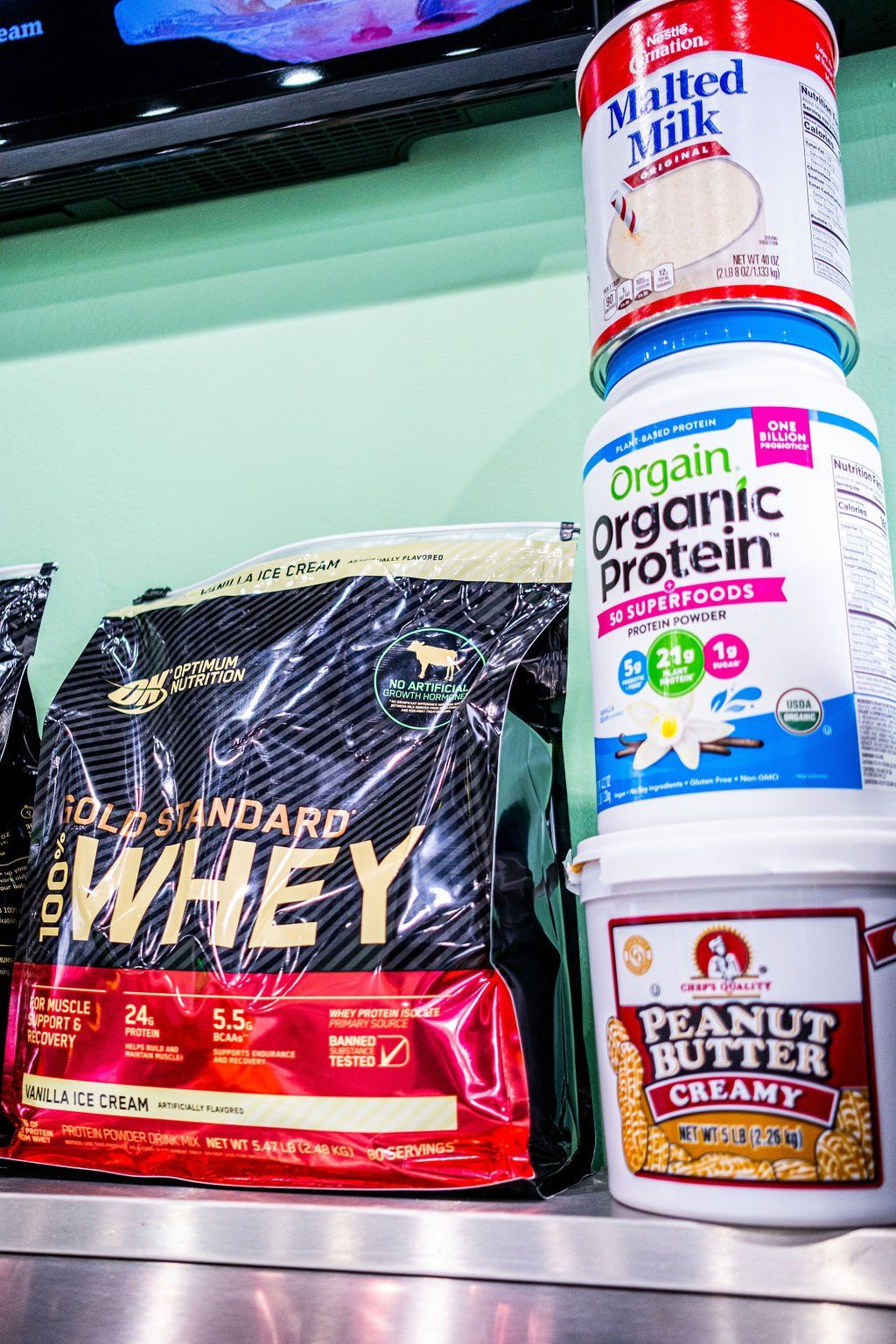 A stack of Orgain organic protein, malted milk, and peanut butter beside a bag of Optimum Nutrition Gold Standard whey.