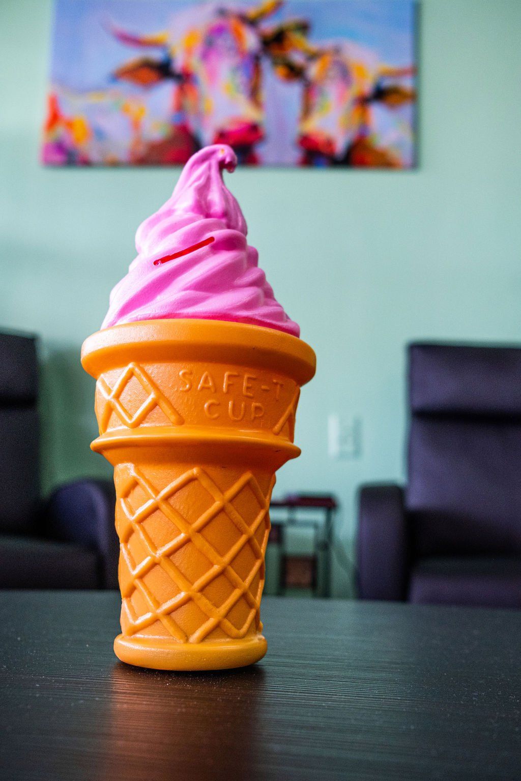 A pink soft-serve ice cream cone-shaped piggy bank sits on a dark table in front of a painting of two cows.