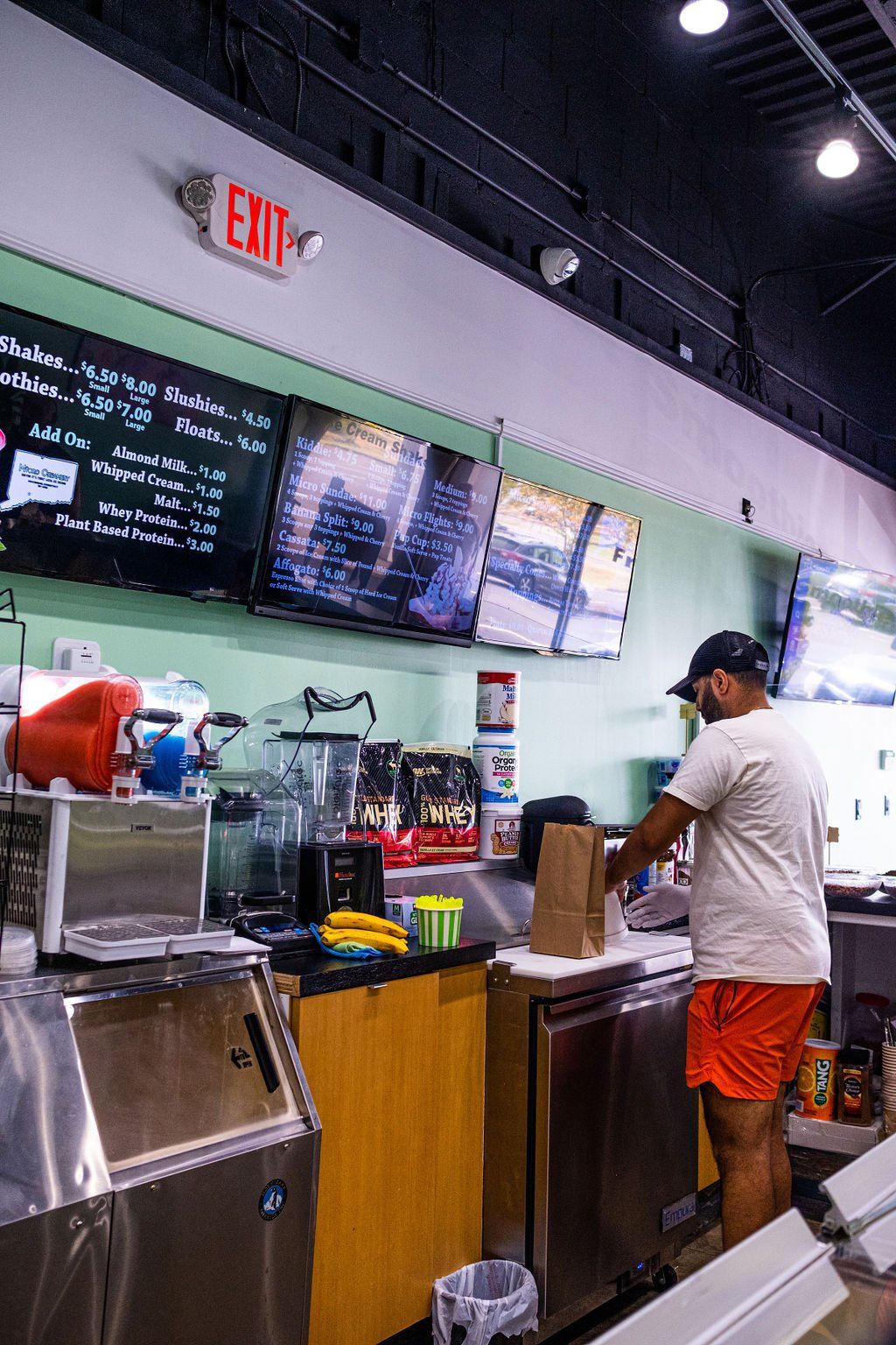 A person in a white t-shirt and orange shorts prepares a brown paper bag behind a counter in a food service setting.