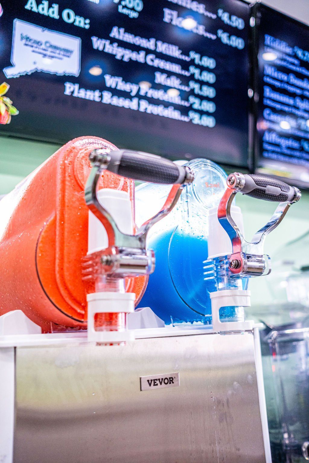 A VEVOR slush machine dispenses bright orange and blue frozen drinks beneath a menu board.