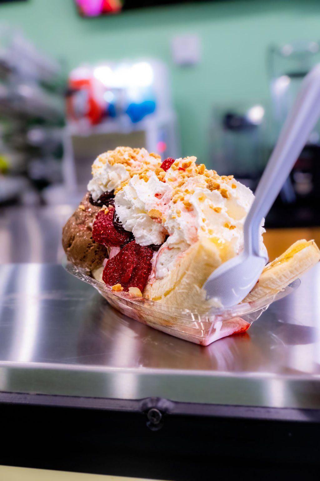 A banana split in a plastic boat on a metal counter, topped with ice cream, whipped cream, strawberries, and nuts.
